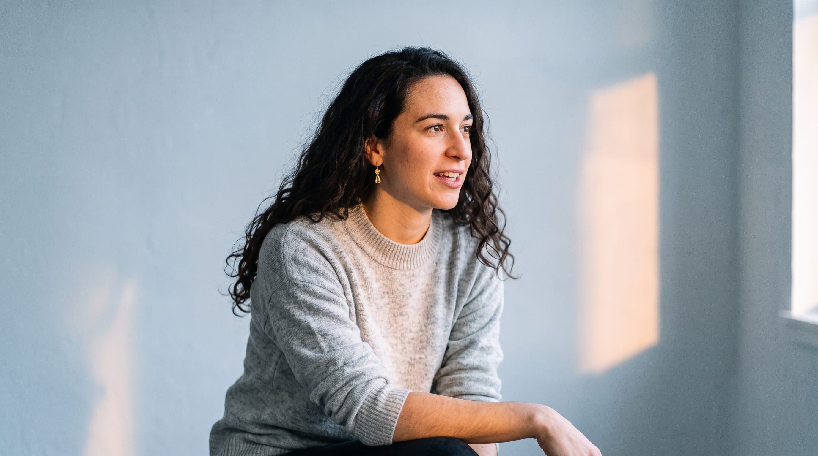 Maya, smiling, seated by a window in a grey sweater.